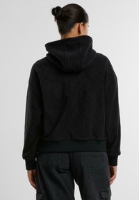 Felpa nera corta con tessuto testurizzato e cappuccio. Presenta polsini e vita a costine. Indossata con pantaloni cargo scuri, vista da dietro.