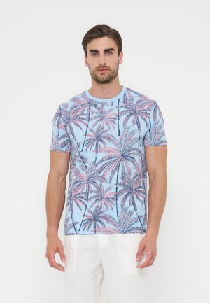 Muž oblečený v svetlomodrom tričku s ružovým a tmavomodrým vzorom paliem a bielych nohaviciach, stojaci pred jednofarebným bielym pozadím.