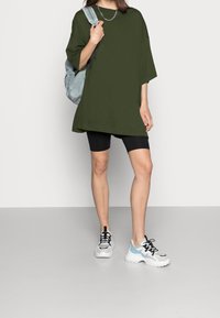 Overwymiarowy oliwkowozielony t-shirt z krótkim rękawem, zestawiony z czarnymi szortami rowerowymi i białymi trampkami z niebieskimi i czarnymi akcentami.