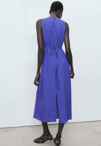 Purple sleeveless dress with black polka dots, cinched waist tie, knee-length split, made of lightweight fabric. Simple and straight cut.