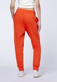 Orange Sweatpant im lockeren Schnitt, mit elastischem Bund, seitlichen Taschen und gerippten Bündchen; kombiniert mit weißen Sneakers vor einem schlichten Hintergrund.