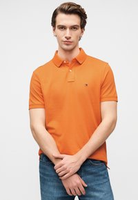 Πορτοκαλί πόλο μπλουζάκι από βαμβάκι με κλασικό γιακά, κοντά μανίκια και μικρό λογότυπο στο στήθος, συνδυασμένο με μπλε τζιν.