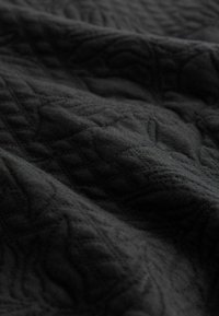 Nahaufnahme von schwarzem Stoff mit erhöhten, strukturierten geometrischen Mustern und subtilen Falten, die Tiefe und Schatten erzeugen.