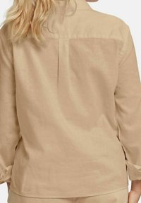 Camicia di lino beige a maniche lunghe, con uno spacco sul retro e una texture sottile. Polsini arrotolati con dettaglio a tab fisso.