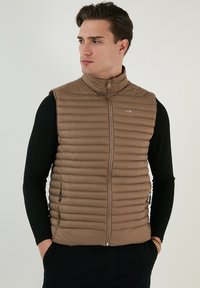 Vest căptușit de culoare maro deschis, cu guler �înalt, textură cu quilt vertical, fermoar frontal și buzunare laterale, purtat peste o cămașă neagră cu mâneci lungi.