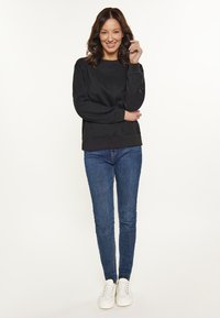 Fekete pulóver, kerek nyakkivágással, kék skinny farmernadrággal és fehér sportcipőkkel párosítva. A modell mosolyogva áll, kezeit a derekán keresztbe téve.