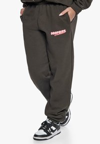 Dunkelgraue Jogginghosen mit elastischem Bund, seitlichen Taschen und pinkfarbenem Logo-Detail. Getragen mit schwarz-weißen Turnschuhen.