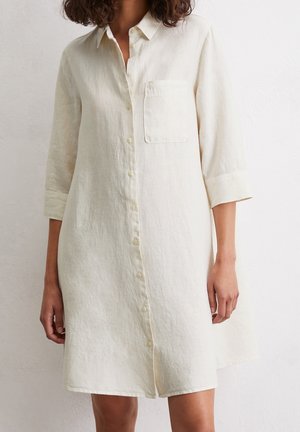 Femme portant une robe boutonnée crème, à manches longues, longueur genou, avec une poche sur la poitrine.