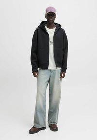 Sudadera negra con cremallera y dos bolsillos frontales, combinada con una camiseta gráfica blanca y vaqueros de pierna ancha de color azul claro. Zapatos marrones de suela gruesa.