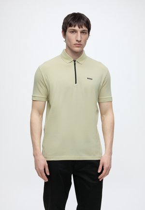 Młody mężczyzna w jasnobeżowej koszulce polo z krótkim rękawem z czarnym zamkiem błyskawicznym i logo "BOSS", w czarnych spodniach, stoi na białym tle.