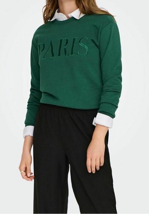 Kobieta w zielonej bluzie z wytłoczonym napisem „PARIS”, na białej koszuli z kołnierzykiem, w połączeniu z czarnymi, wysokimi spodniami.