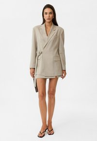 Beige blazer jurk met een op maat gemaakte ontwerpen, enkelvoudige sluiting en zijstrikdetail. Heeft lange mouwen en een gestructureerde pasvorm.