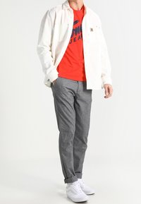 Biała koszula zapinana na guziki noszona na czerwonym t-shircie z niebieskim napisem, szare wzorzyste spodnie i białe trampki. Model stoi na neutralnym tle.