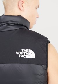 Gilet senza maniche nero realizzato in materiale imbottito. Presenta un colletto alto e un logo ricamato bianco sul lato sinistro. Superficie liscia.
