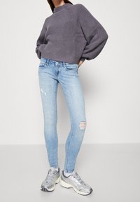 Harmaa neulottu neulepusero korkealla kauluksella, yhdistetty vaaleansiniin kapealahkeisiin farkkuihin, joissa on kulutetut yksityiskohdat, ja harmaisiin urheilutossuihin.