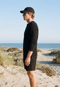 Uomo con cappello nero, occhiali da sole, camicia a maniche lunghe e pantaloncini in piedi su una spiaggia sabbiosa con il mare e il cielo sullo sfondo.