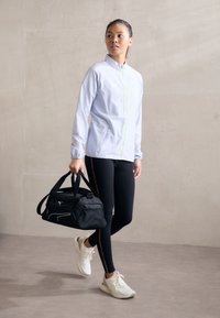 Lysblå træningsjakke med en grøn lynlås, sorte leggings med sidelister og hvide sneakers, mens der bæres en sort duffeltaske.