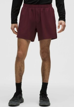 Männlich, trägt dunkelrote Sportshorts, ein schwarzes langärmeliges Oberteil, schwarze Socken und schwarze Laufschuhe vor einem einfarbigen Hintergrund.