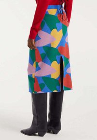 Persona vestida con una falda colorida con formas abstractas, una blusa roja y botas negras hasta la rodilla, de pie contra un fondo simple.