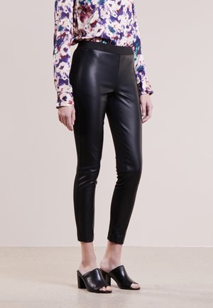 Femme portant un pantalon en cuir noir ajusté, un chemisier multicolore à manches longues et des mules noires à talons ouverts, se tenant devant un fond uni.