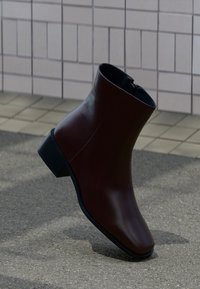 Bottine en cuir marron foncé avec un petit talon bloc, fermeture éclair latérale, sur un sol carrelé avec un arrière-plan de pavés gris.