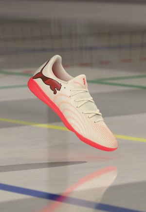 Chaussure de football en salle blanche et rouge avec logo Puma noir, flottant au-dessus d’un sol de gym visé par des lignes de terrain colorées.
