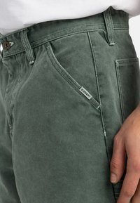 Pantalons en denim verts avec une coupe droite, des coutures marquées et une petite étiquette de marque sur la poche avant.