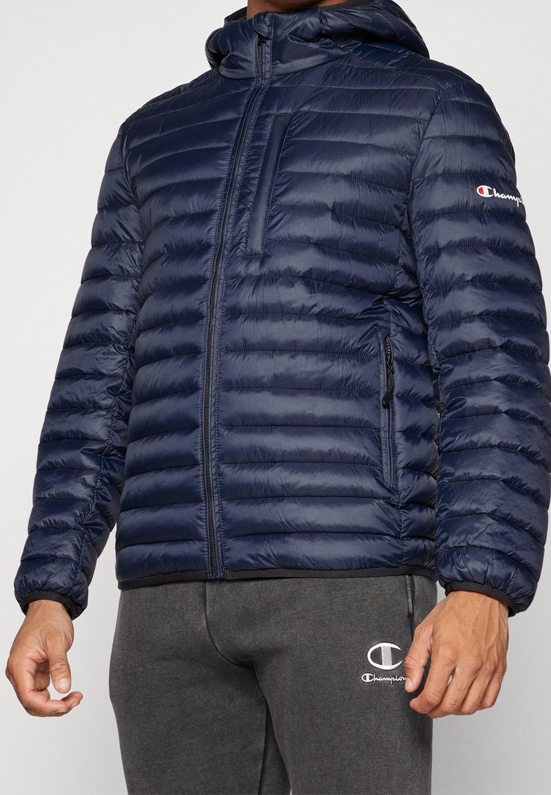 Navy puffer jacket with horizontal stitched pattern, full zip, attached hood, and side pockets. Features Champion logo on the left sleeve.