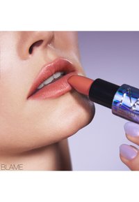 Un tube de rouge à lèvres nude avec un étui métallique irisé appliqué sur des lèvres lisses et légèrement brillantes. Le teint est chaud, avec des dents visibles.