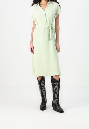 Femme portant une robe midi ceinturée vert clair à manches courtes et des bottes noires montantes avec des détails brodés blancs.