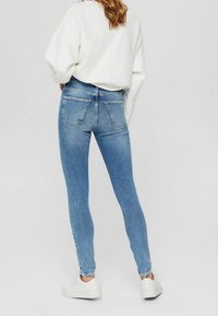 Jeans de denim azul ajustados con dos bolsillos traseros, diseño de cintura alta y una sutil textura desgastada. Llevados con un suéter blanco holgado y zapatillas blancas.