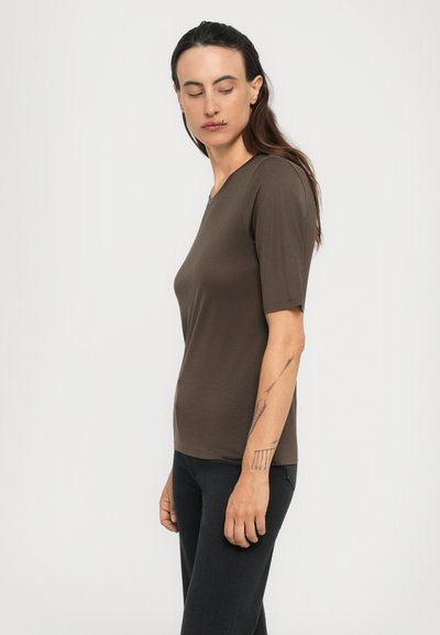Donna con lunghi capelli scuri che indossa una camicia marrone a maniche corte e pantaloni neri, che guarda in basso e di lato su uno sfondo neutro.