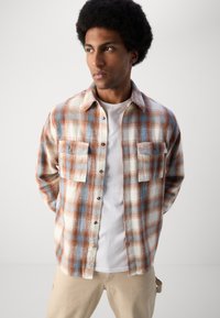 Plaid flannel shirt in cream, orange, and blue with two chest pockets and button closure. Worn over a white T-shirt. Lightweight fabric.