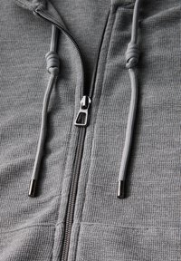 Grauer Reißverschluss-Hoodie aus strukturiertem Stoff, mit verstellbaren Kordeln und metallischem Reißverschluss. Verfügt über eine glatte Oberfläche und praktisches Design.