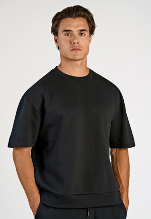 Zwarte korte mouwen trui gemaakt van een zachte, gladde stof. Voorzien van een ronde hals en een losse pasvorm, met zichtbare stiksels langs de randen.