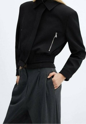 Persoon die een zwart cropped jasje draagt met een voorzak met rits, gecombineerd met hooggetailleerde, geplooide, donkergrijze, wijde pijpen broek.