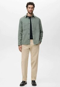 Hellgrünes Hemd mit zwei Brusttaschen, schwarzes T-Shirt mit Rundhalsausschnitt darunter, beige Hose und schwarze Slipper. Männliches Model steht vor einem weißen Hintergrund.