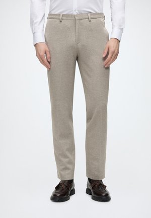 Homme portant un pantalon beige sur mesure, une chemise blanche et des mocassins en cuir marron foncé à glands, debout, les mains détendues.
