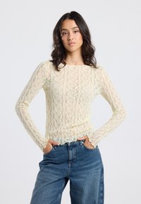 Woman with wavy dark hair wearing a sheer cream lace long-sleeve top and blue high-waisted jeans, standing with hands in pockets.