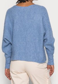 Hellblauer, lockerer Strickpullover mit ribbelten Ärmelbündchen und strukturiertem Muster. Das Rückendesign ist etwas länger als die Vorderseite.