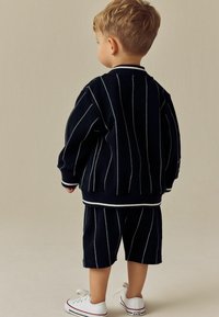 Marineblauw gebreid trainingspak met witte verticale strepen, geribbelde manchetten en shorts. Gecombineerd met witte sneakers met rode accenten.