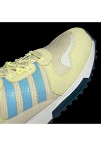 Zapatilla deportiva de color amarillo claro que presenta una tela de malla, acentos de rayas azules, superficie texturizada, detalles en blanco y una suela de goma negra.