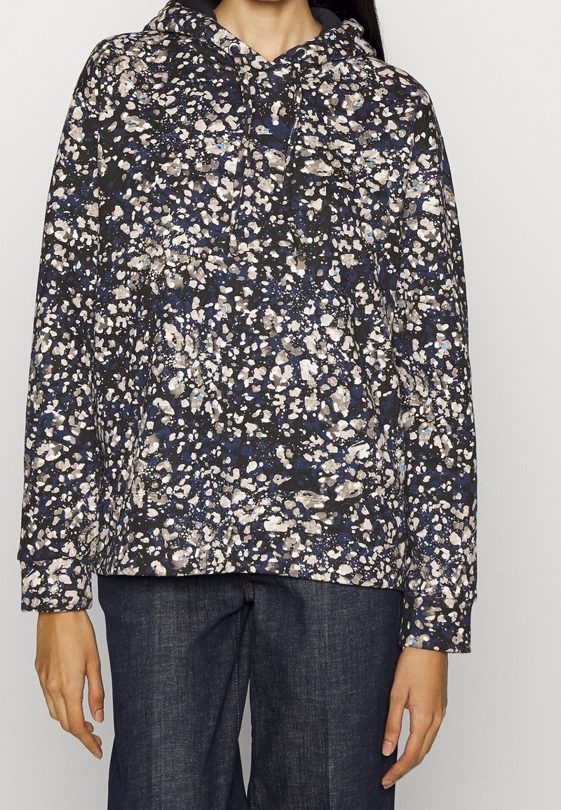 Femme portant un sweat à capuche noir avec un imprimé floral abstrait blanc et bleu et un jean bleu foncé.