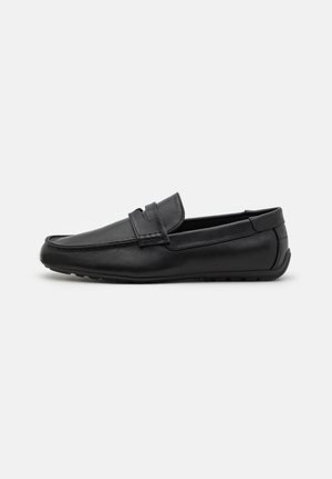 Mocassin en cuir noir avec lanière penny, couture moc toe, et semelle en caoutchouc, présenté en vue latérale sur un fond uni.