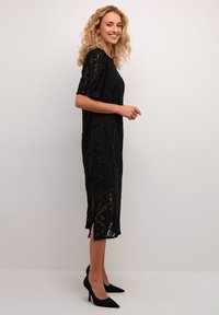 Black lace midi dress with short sleeves, floral pattern, side slits, and a relaxed fit, paired with pointed black heels.