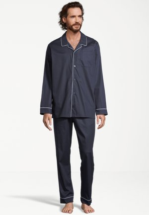 Mann steht barfuß und trägt dunkelblaue langärmelige Pyjamas mit weißen Paspeln und einem Hemd mit Knöpfen vor einem schlichten weißen Hintergrund.