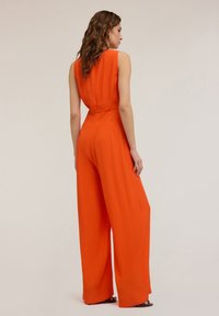 Tuta arancione senza maniche con design a gamba larga. Tessuto liscio, dettagli sotto cucitura sul retro e corpino aderente. Stile minimalista.