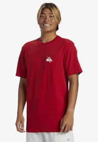 Camiseta de algodón roja con mangas cortas y cuello redondo. Presenta un pequeño gráfico blanco en el lado izquierdo del pecho. Textura suave y corte relajado.
