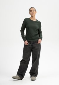 Donkergroene langemouwen shirt met ronde hals, gecombineerd met losse zwarte jeans en grijze instappers. Gladweefsel textuur.