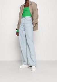 Person som bär en beige kavaj över en grön topp, ljusblå vida jeans och vita sneakers, stående mot en plain vit bakgrund.